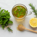 Refreshing Peppermint Tea Recipe for Mental Clarity 2 A flat lay of ingredients for a fresh peppermint tea recipe, including a bunch of peppermint, a sprig of rosemary, a lemon slice, and a honey dipper next to a clear glass mug of the finished tea.