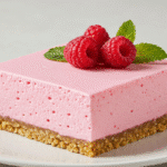 Easy No-Bake Keto Jello Cream Cheese Dessert 2 A square slice of pink raspberry **keto jello cream cheese dessert** with a nut crust, served on a white plate and garnished with fresh raspberries and mint.