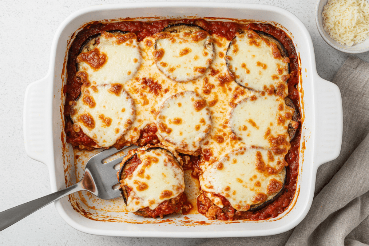 Easy Baked Eggplant Parmesan Casserole An overhead shot of a finished baked eggplant parmesan calories in a white ceramic baking dish, with one cheesy slice being lifted out.