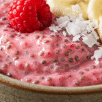 Chia and Raspberry Pudding That Will Transform Your Mornings 2 An extreme close-up of a chia and raspberry pudding in a rustic bowl, topped with a fresh raspberry, banana slices, and shredded coconut.
