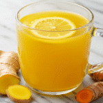 Soothing Lemon Ginger and Turmeric Tea Recipe 2 A close-up of a finished lemon ginger and turmeric tea in a clear glass mug, with fresh ginger and turmeric root on a white marble surface.
