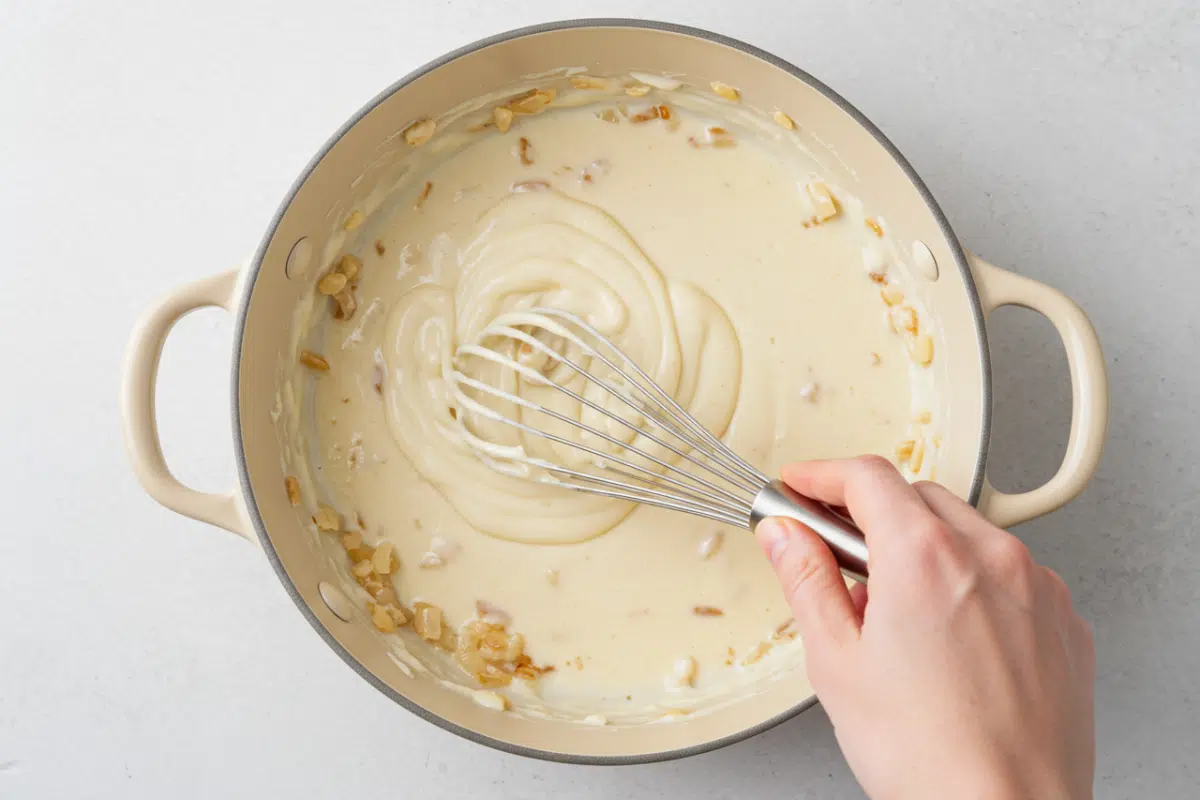 How to Make a Creamy Sauce for Pasta Bake A hand whisking a smooth, creamy cheese sauce in a light-colored pan for a chicken broccoli pasta bake.