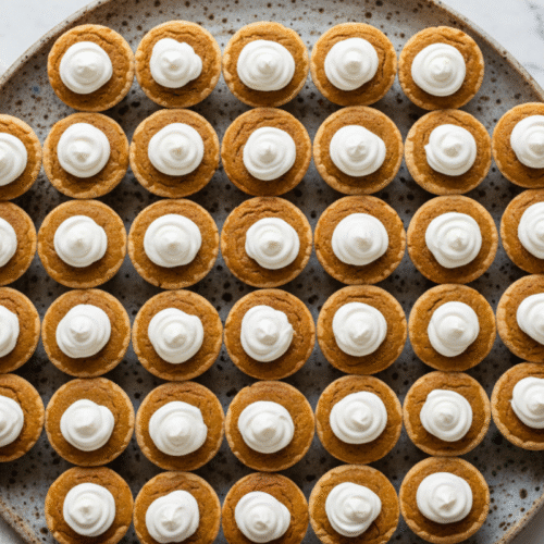 Overhead view of a large platter of mini pumpkin pie recipe bites with whipped cream topping.