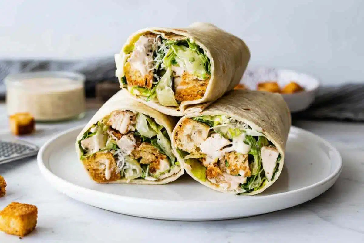 Easy Chicken Caesar Wrap 1 Easy chicken Caesar wrap recipe showing three stacked wraps on white plate with cross-section of romaine lettuce, grilled chicken, croutons and Caesar dressing