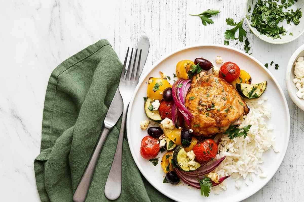 Greek Sheet Pan Chicken Recipe Greek sheet pan chicken served on white plate with roasted bell peppers, cherry tomatoes, red onions, Kalamata olives, crumbled feta cheese, and fresh parsley garnish alongside white rice